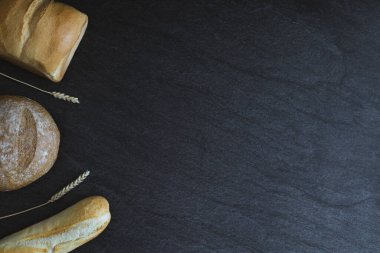 Two loaves and one French baguette with ears lie on the left on a black stone background with copy space on the right, flat lay close-up. Bread baking concept.
