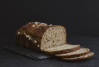 One rye dark bread with crushed oat grains and sunflower seeds lies cut into thin pieces lies on a stone stand on the left on a black background, flat lay close-up.The concept of baking bread.