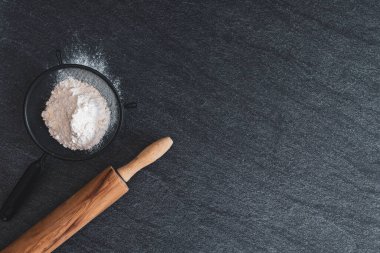 One sieve with flour and a wooden rolling pin lie on the left on a black stone table with copy space on the right, flat lay close-up. The concept of baking bread.