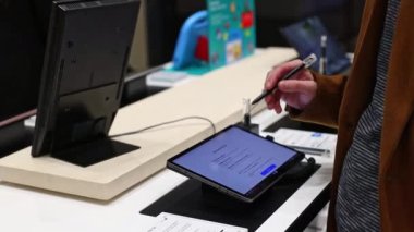 A young Caucasian unrecognizable man in a brown coat inspects a new tablet, checking how the stylus works by swiping it across the screen, standing behind the counter of the store, close-up from the side in slow motion,. Smartphone shopping concept.