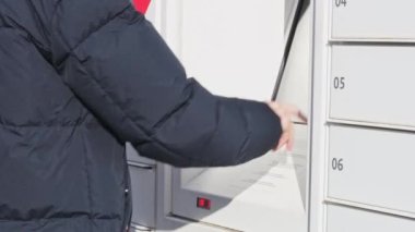 A young caucasian man in a blue jacket enters a code with his finger on the screen of a mailbox to take a parcel, close-up side view. The concept of receiving a parcel, a modern lifestyle.