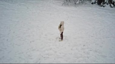 A young caucasian girl in a white hooded down jacket and brown pants stands in the winter forest of a nature reserve in belgium, side view close-up with circular tracking from a drone. Winter holiday concept, modern lifestyle.