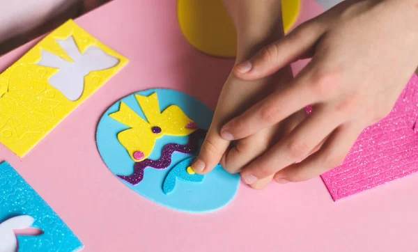 Mother's hands help a little caucasian girl stick with her fingers a yellow circle of felt sticker on a blue egg with a hat and a lilac zigzag, sitting at a children's table with a set of craft predas on a pink background with depth of field, close-u