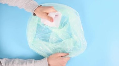 Genç beyaz bir adamın elleri beyaz plastik bardakları sıkar ve mavi arka planda plastik bir torbaya koyar..