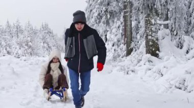 A young caucasian man rides a sitting girl on a sled in the winter forest of a nature reserve in belgium, close-up side view. The concept of winter holidays, modern lifestyle.