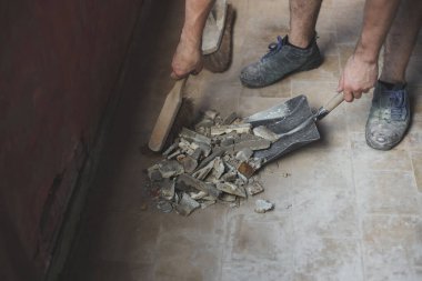 Kirli fayanslı bir zeminden eski bir faraş üzerinde küçük bir çırpıyla inşaat enkazını süpüren tanınmayan genç beyaz bir adam. Pencereleri temizleme ve kurma konsepti, inşaat işi, ev yenileme.