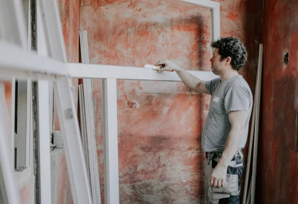 Young caucasian man holding a yellow construction knife and cleaning a dirty window frame while standing against a red wall, close-up side view. The concept of cleaning and installing windows, construction work, house renovation.