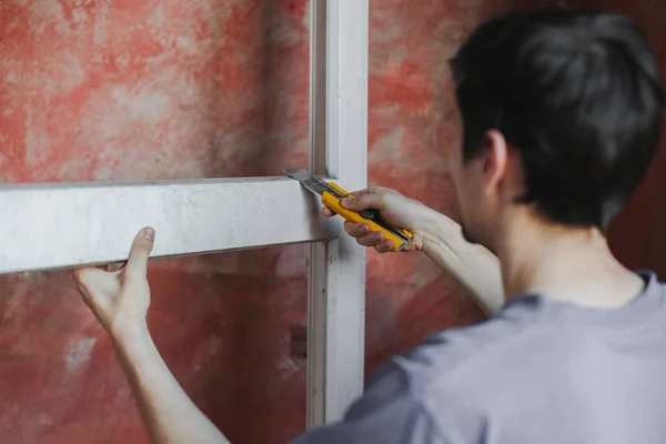 Young caucasian man cleans a dirty window frame with a yellow construction knife holding it with his hand against the background of a red wall, close-up side view. The concept of cleaning and installing windows, construction work, house renovation.