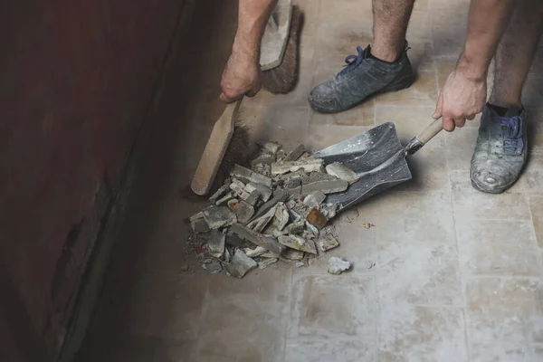 Young caucasian unrecognizable man sweeping construction debris with a small whisk on an old dustpan from a dirty tiled floor, close-up side view. The concept of cleaning and installing windows, construction work, house renovation.