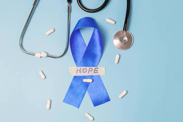 One blue ribbon with sticky tape with the word: hope, stethoscope and scattered pills on a blue background, flat lay close-up. World cancer day concept, abdominal diseases.