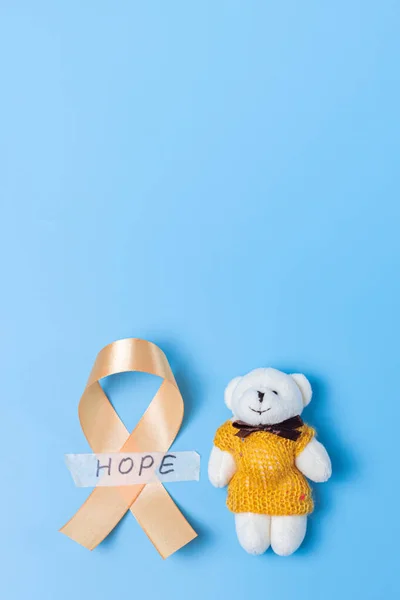One yellow ribbon with sticky tape with hope and a toy bear lie from below on a blue background with copy space from above, flat lay close-up.Concept for World Childhood Cancer Day.