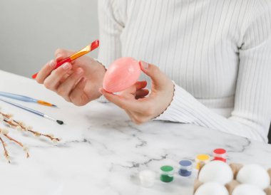 Hands of a caucasian teenage girl in a white turtleneck holding an egg and painting with a pastel pink acrylic brush while sitting at a marble table for diy preparation for the easter holiday, close-up side view. The concept of crafts, needlework, ar