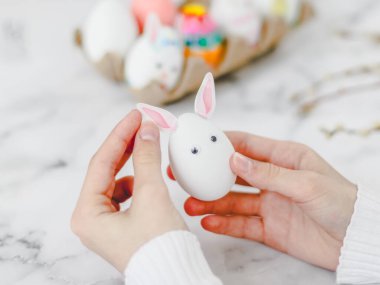 Çocukların elleri beyaz bir yumurtayı tutarken gözleri yapış yapış, kulakları tavşan kulaklarıyla yapıştırır, mermer bir masaya oturur, Paskalya bayramı için el sanatları hazırlar, yakın plan yan görüş için. Zanaat konsepti, dikiş işi, evde, diy, çocuk sanatı,