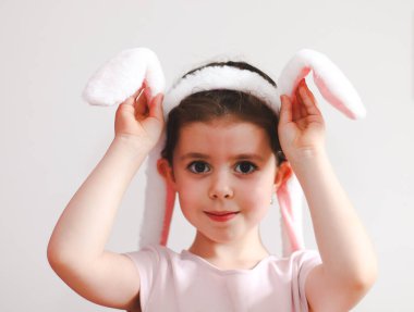 Pembe tişörtlü güzel beyaz bir kız kameraya bakıp gülümseyerek ellerini tutuyor ve beyaz arka planda tavşan kulaklığını hareket ettiriyor. Kavram, pankart, kurusıkı, tatil, mutlu paskalya, çocuklar