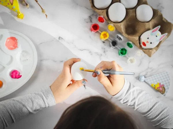 stock image Hands of a caucasian girl in a gray turtleneck, paint an easter egg with a brush and acrylic paint on a marble table with a palette, willow branches, eggs in a box for diy preparation for the easter holiday, flat lay closeup. The concept of crafts, n