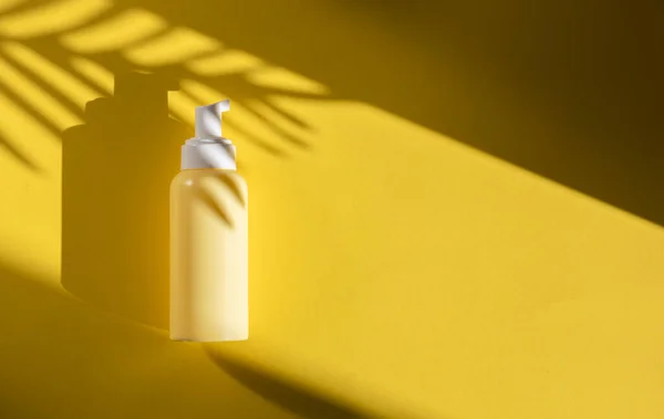 One small yellow bottle with a dispenser and the shadow of a palm tree branch lies on the left against a yellow background with hard shadows from the sun and copy space on the right, flat lay close-up. Cosmetics concept.