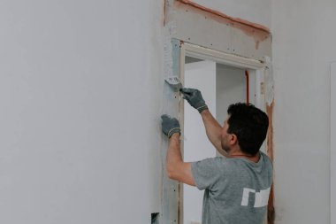 Gri üniforma ve eldivenlerle tanınan genç beyaz bir beyaz adam, kapı aralığında yarım bir dönüş yaparken, seçici bir odak ile bina ağını beyaz macunla kaplıyor. Ev yenileme konsepti, inşaat işi.