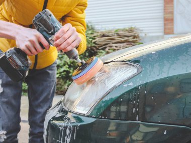 Kafkasyalı genç bir adamın elleri, süngerli bir matkapla matkabı deliyor ve arabasının farını deterjan sabunuyla parlatıyor. Yakın plan, araba yıkama ve araba farlarıyla temizlik kavramı..