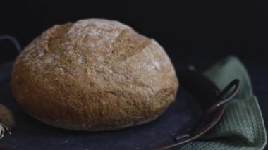 Kulakları ve zeytinli mutfak peçetesi olan klasik metal bir tepside bir yuvarlak çavdar ekmeği siyah taştan bir arka planda duruyor, yakın plan solda iz bırakarak. Ekmek pişirme konsepti.