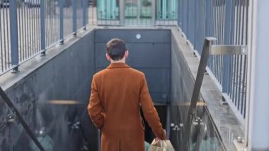 Arkadan kahverengi paltolu, elinde paketler olan yakışıklı, beyaz tenli, esmer bir adam metroya doğru iniyor. Çevrimdışı alışveriş kavramı, erkek yaşam tarzı..