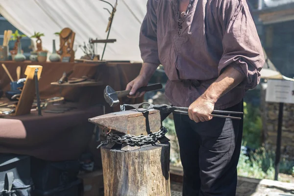 One middle-aged caucasian male blacksmith forges iron with a sledgehammer, standing behind a wooden stump on the street of Luxembourg city medieval castle, side view, close-up. The concept of medieval needlework, artisanal.