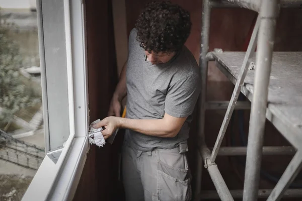 A young caucasian man standing at the window wipes a metal spare part on the window frame in the room where the repair is underway with a paper napkin, close-up side view with selective focus. The concept of home renovation, washing window frames.