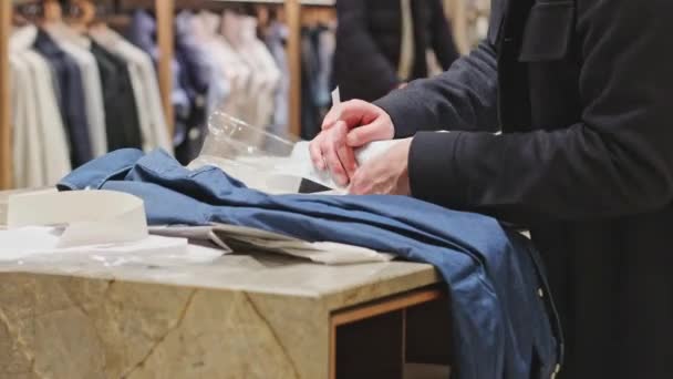 年轻的高加索推销员把一件折叠的蓝色夹克整齐地包在白纸里 用胶带粘在一起 站在柜台后面的时尚服装店里 近距离观察 离线购物概念 现代生活方式 — 图库视频影像