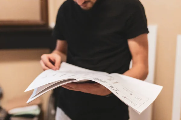 A young caucasian guy holds a paper instruction book on assembling furniture in his hands and carefully reads, side view close-up with depth of field.