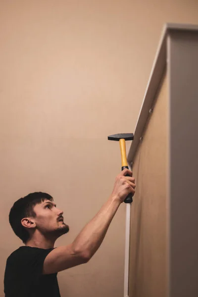 Young caucasian brunette man hammers with a yellow mallet plastic rivets in the back plywood wall of the wardrobe in the bedroom, close-up side view with selective focus. Furniture assembly concept.