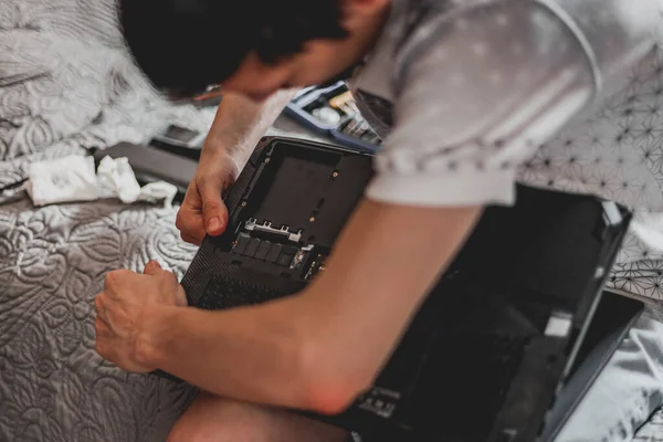 Young caucasian unrecognizable guy connects spare parts in a laptop, closing it after repair, sitting on a bed in a room, close-up side view. Step by step concept of laptop repair and thermal paste replacement.