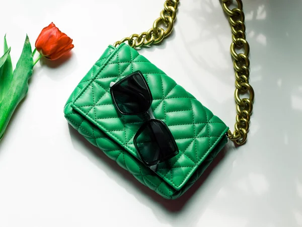 Stylish women's trendy green quilted bag with a large gold chain, a red tulip in it and sunglasses lie on a white table with shadows from the window, flat lay close-up. Fashionable accessories concept.