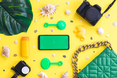 İki kamera, yeşil bir el çantası, bir canavar yaprağı, kum için hazırlanmış bir çocuk, deniz kabukları ve ortasında yeşil ekran olan bir akıllı telefon, fotokopi alanı olan sarı bir arka plan, düz yakın plan. Yaz konsepti, yaz blogcusu, yaz zamanı.