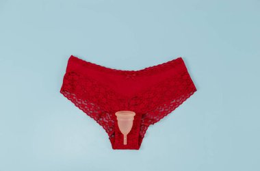 Silikon regl fincanı olan kırmızı dantelli bir kadın külotu mavi arka planda, düz bir zeminde yatıyor. Kadın sağlığı kavramı.