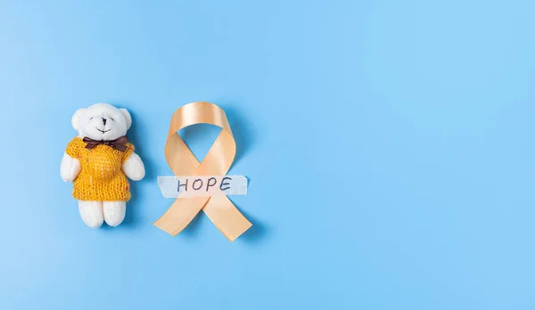 One yellow ribbon with sticky tape with hope and a toy bear lie on the left on a blue background with copy space on the right,flat lay close-up.Concept for World Childhood Cancer Day.