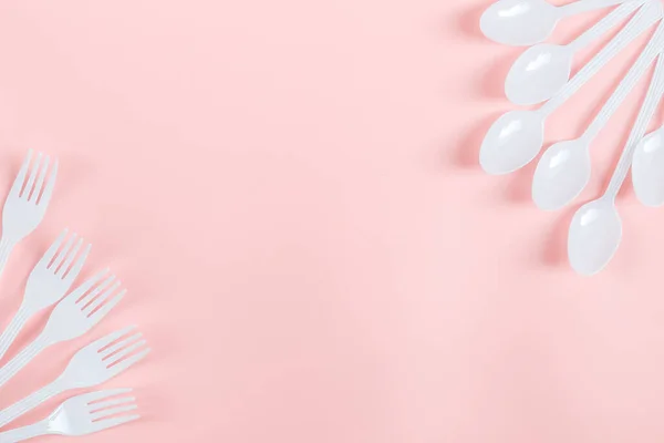 White plastic forks and spoons lie on the sides on a light pink background with copy space for text in the center, flat close-up. The concept of ecology, plastic garbage and disposable plastic tableware.