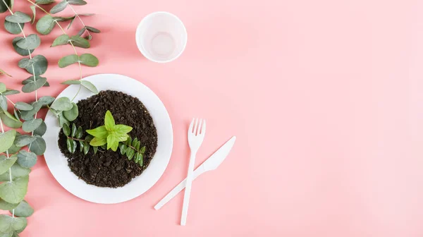 stock image White plastic cup, crossed fork and knife, plastic plate with black soil and green sprouts on the left on a light pink background with copy space for text on the right, flat lay close-up. The concept of ecology, plastic garbage and disposable plastic