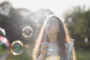 Gri uzun kollu tişörtlü, kahverengi uzun saçlı beyaz bir kızın bulanık portresi. Parktaki bir çayırda sabun köpükleri üflüyor. Arka planda bir oyun parkında, seçmeli odak ile yakın plan. Park ve REC kavramı, mutlu çocukluk, masal.