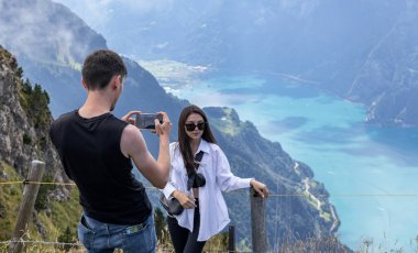 Arkadan genç beyaz bir adam cep telefonuyla güzel kız arkadaşının fotoğraflarını çekiyor. Fronalpstock İsviçre 'de göllerin ve dağların arka planında, yakın çekimde..