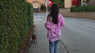 Arkadan pembe gömlekli ve cep telefonlu genç beyaz bir kız şehirde gece vakti tasmalı minyatür bir pinscher ile yürüyor. Köpek gezdirme konsepti..