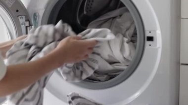 Genç, beyaz, tanınmayan bir kız yerde otururken çizgili yastık kılıflarını çamaşır makinesine koyuyor. Çamaşır yıkama konsepti.