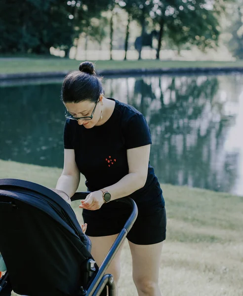 Caucasian young and beautiful woman mother in a black t-shirt and shorts pulls her hands to her little daughter sitting in a stroller from the back in a public park on the lawn near the lake, close-up