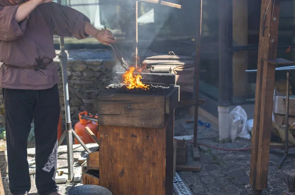 One middle-aged caucasian male blacksmith melts iron in fire holding it with tongs, standing behind a wooden stand on a Luxembourg city street in a medieval castle side view, close-up. The concept of medieval needlework, artisanal.