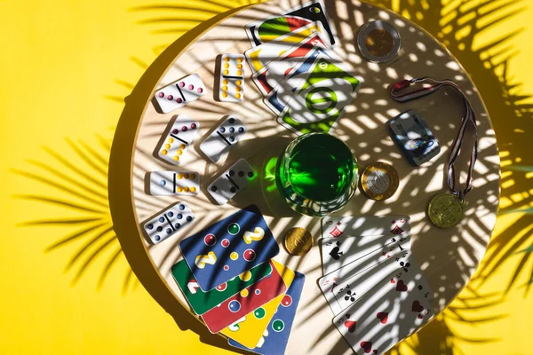 白色的多米诺骨牌 扑克牌 单簧管 棒棒糖 巧克力硬币 获奖者奖章和绿色饮料在一个黄色背景的圆形木制旋转板上的杯子里 有一个棕榈树枝的影子 平平平静静地躺着 概念夏季棋盘游戏 — 图库照片