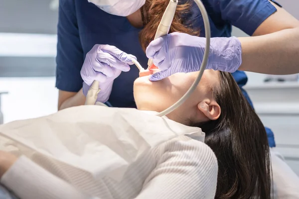 A dentist in a blue uniform and lilac gloves cleans the gums and teeth with a dental electric hook in the oral cavity of a young brunette girl patient lying in a chair, close-up view from below. Concept, health and oral hygiene.