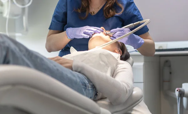 A dentist in a blue uniform and lilac gloves polishes and whitens teeth using dental tools of a young brunette patient lying in a chair, close-up view from below. Concept, health and oral hygiene.