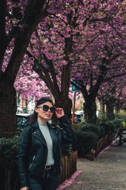 Güneş gözlüklü ve deri ceketli güzel beyaz bir kızın portresi şehrin arka planında duruyor. Sakura ağaçlarının çiçek açmasına karşı..