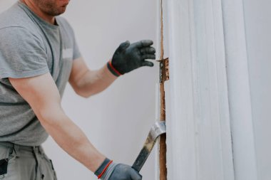 Üniformalı ve gri tekstil eldivenli genç beyaz adam, levye kullanarak eski ahşap şeritleri kapı aralığından kaldırıyor..