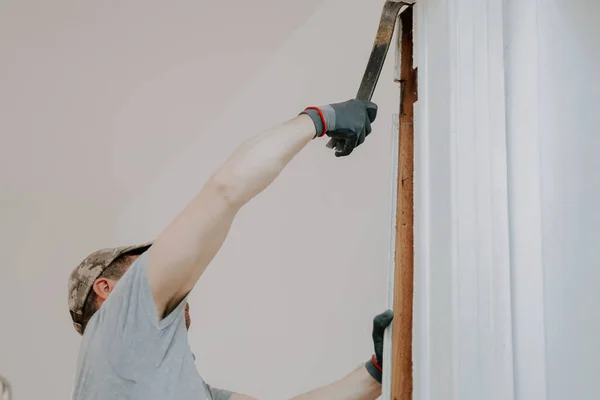 stock image Young caucasian man in uniform and gray textile gloves using a crowbar removes old wooden plinths from a doorway, bottom view close-up with selective focus.Construction work concept.