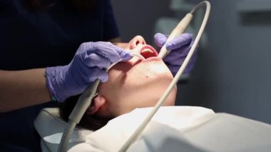 Leylak rengi eldivenli genç bir dişçinin elleri diş matkabı ve suyla kadın bir hastanın ağız boşluğunu temizler..