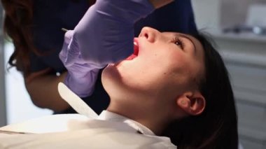 Mavi üniformalı, leylak eldivenli bir dişçi, sandalyeye uzanmış, yan görüntüsü olan genç ve güzel bir kızın ağız boşluğuna anestezi enjekte eder..